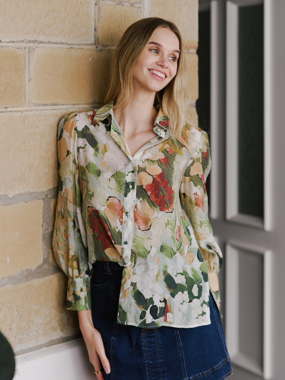 A woman standing against a wall, wearing a floral print blouse with full sleeves and a collar, paired with blue denim shorts.